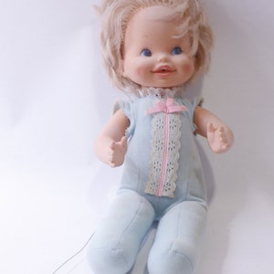 May include: A vintage pull-string doll with blonde hair, blue eyes, and a pink bow. The doll is wearing a light blue sleeper with white lace trim.