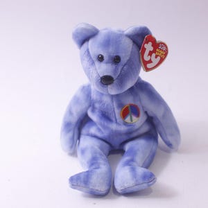 Könnte beinhalten: Ein flauschiger, lavendelfarbener Teddybär mit einem Friedenszeichen auf der Brust. Der Bär hat schwarze Augen und eine schwarze Nase. Ein rotes herzförmiges Etikett ist an seinem Ohr befestigt.
