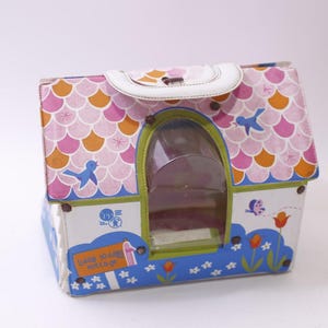 May include: A vintage toy house-shaped carrying case with a white handle. The roof is patterned with pink, orange, and white scales. The house has a clear plastic window and is decorated with blue birds, flowers, and the text "Happy 10 Years Cottage".