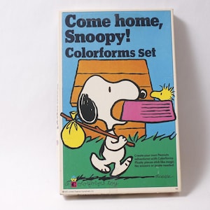 May include: A vintage Colorforms set box featuring Snoopy from the Peanuts comic strip. The box has a blue background with the text "Come home, Snoopy! Colorforms set." Snoopy is depicted carrying a bundle and a pink object. The box is rectangular.
