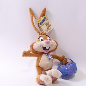 Peut inclure: Une peluche représentant un lapin brun avec de longues oreilles, un nez rose et un nœud papillon rouge. Le lapin tient un panier bleu rempli d'œufs colorés et porte une étiquette Nesquik. Le jouet est conçu pour ressembler au lapin Nesquik.