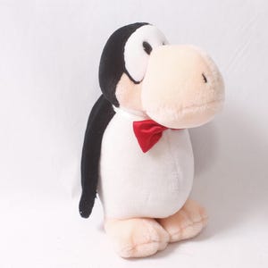 Pode incluir: Um brinquedo de pelúcia de pinguim com cabeça e asas pretas, corpo branco e bico e pés bege claro. Possui uma gravata borboleta vermelha. O pinguim está em pé sobre uma superfície branca.
