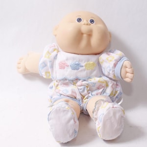 May include: A soft-bodied doll with a round, pale vinyl head, blue eyes, and a small nose. The doll wears a white romper featuring a pastel-coloured pattern of small shapes and three embroidered sheep in pink, blue, and yellow on the chest.