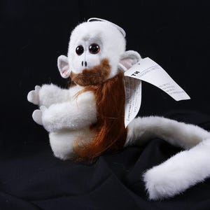 May include: A plush monkey toy with white fur, large dark eyes, and a brown beard. The toy features long, straight reddish-brown hair on its head and back, a long white tail, and a white paper tag attached to its side with visible text.