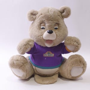 May include: A plush, tan-colored teddy bear with a purple shirt and the text "TV Teddy". The bear has a black nose, open mouth, and white accents on its paws. It is sitting with its arms raised.