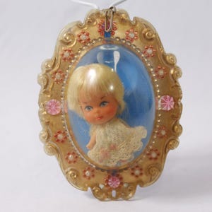 May include: Vintage pendant featuring a doll inside an oval frame. The frame is gold-colored with floral and pink gem accents. The doll has blonde hair and is wearing a white dress. The background is blue, and the pendant hangs from a white cord.