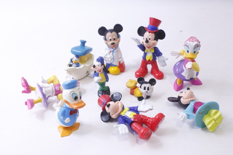 May include: A collection of Disney character figurines, including Mickey Mouse, Donald Duck, and Goofy. The figures are brightly colored and feature classic designs. Some are standing, while others are lying down.