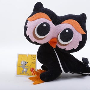 May include: A plush owl toy with a black body, pink face, and orange accents. The owl has large black eyes and a tag with a cartoon illustration. The toy is made of soft fabric.