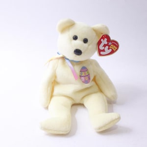 May include: A cream-colored plush teddy bear with black eyes and a black nose. The bear has a colorful Easter egg design on its chest and a red heart-shaped tag. A blue ribbon is tied around its neck.