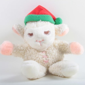 May include: A plush lamb toy with a red and green elf hat. The lamb has closed eyes, pink accents on its ears and paws, and a pink stitched mouth. The lamb is sitting with its arms outstretched.