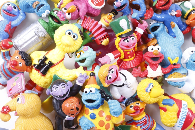 May include: A collection of colorful Sesame Street character figurines, including Big Bird, Elmo, Cookie Monster, and others. The toys are made of plastic and are in various poses, creating a playful and nostalgic scene.
