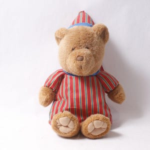 Op de afbeelding: Een bruine teddybeer met een rood-wit gestreepte pyjamaset en een bijpassende nachtmuts met blauwe rand. De beer heeft een zacht, knuffelbaar uiterlijk, perfect voor een kinderspeelgoed of kinderkamerdecoratie.