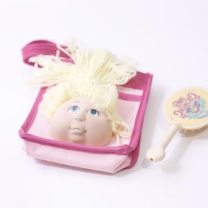 May include: A vintage doll face with blonde yarn hair is set in a pink fabric bag with a magenta trim. A matching yellow and white brush with the text "Growing Hair" is next to the bag. The items are on a white background.