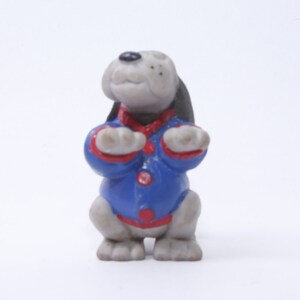 May include: A small gray and white plastic figurine of a dog wearing a blue and red jacket. The dog is standing on all fours with its paws raised in a begging position.
