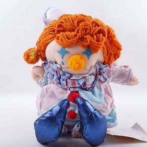 Op de afbeelding: Een pluche clown met oranje garenhaar, blauwe ogen met sterren, een gele neus en een roze-blauwe gepatroneerde outfit. De clown zit met zijn handen voor zich gevouwen.
