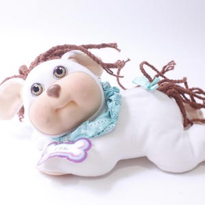 Puede incluir: Juguete de peluche blanco en forma de perro con pelo de lana marrón, ojos marrones y una bufanda con estampado azul y blanco. El juguete tiene una etiqueta rosa con el texto "CPK" en ella.