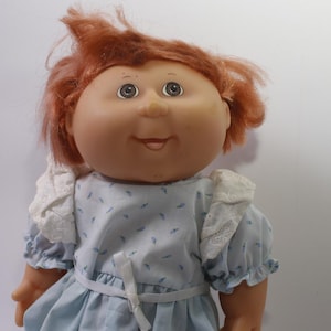 Puede incluir: Muñeca vintage con cara redonda, ojos azules y pelo rojo. La muñeca lleva un vestido azul claro con cuello de encaje blanco y cinturón de cinta blanca. La piel de la muñeca es de color melocotón claro.