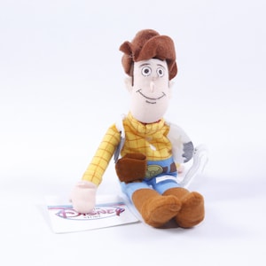 Może przedstawiać: Pluszowa zabawka Woody'ego z serii Toy Story. Woody ma na sobie sw&oacute;j charakterystyczny brązowy kowbojski kapelusz, ż&oacute;łtą kratowaną koszulę, niebieskie dżinsy i brązowe buty. Siedzi z uśmiechem na twarzy. Zabawka ma metkę z napisem "The Disney Store".