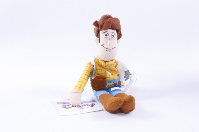 Może przedstawiać: Pluszowa zabawka Woody'ego z serii Toy Story. Woody ma na sobie sw&oacute;j charakterystyczny brązowy kowbojski kapelusz, ż&oacute;łtą kratowaną koszulę, niebieskie dżinsy i brązowe buty. Siedzi z uśmiechem na twarzy. Zabawka ma metkę z napisem "The Disney Store".