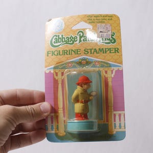 Peut inclure: Un tampon figurine Cabbage Patch Kids vintage dans son emballage d'origine. La figurine représente un personnage portant un manteau jaune, un chapeau rouge et des bottes rouges, tenant une loupe. L'emballage est coloré avec les mots "Cabbage Patch Kids" et "Figurine Stamper".