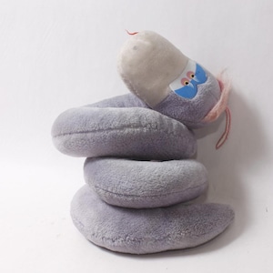 Puede incluir: Un juguete de peluche con forma de serpiente enroscada con pelaje gris suave. La serpiente presenta una cabeza gris claro, parches azules alrededor de los ojos naranjas y un pequeño mechón de pelo rosa en la parte posterior de la cabeza.