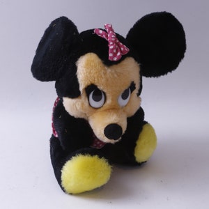 May include: A plush toy of a cartoon mouse with black ears, a yellow face, and a red bow with white polka dots. The toy is sitting with its legs crossed and has large, black eyes.