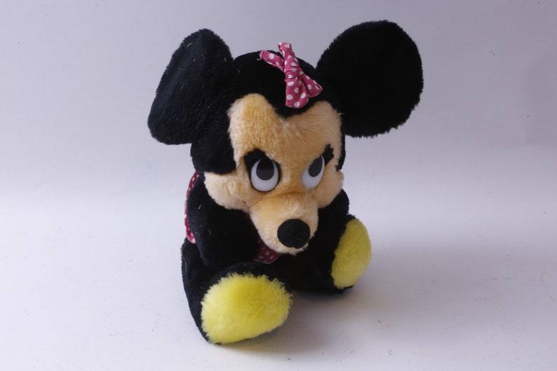 May include: A plush toy of a cartoon mouse with black ears, a yellow face, and a red bow with white polka dots. The toy is sitting with its legs crossed and has large, black eyes.