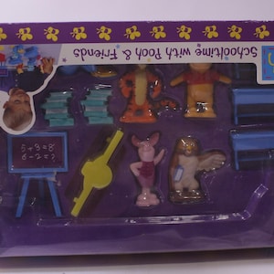 May include: A boxed playset featuring Winnie the Pooh and Friends. The set includes figurines of Pooh, Tigger, Piglet, and Owl, along with school-themed accessories like desks, books, and a chalkboard. The packaging is purple and white.
