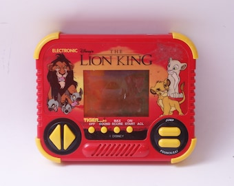 Op de afbeelding: Een rood en geel handheld elektronisch spel met het thema The Lion King van Disney. Het spel heeft een scherm, knoppen en afbeeldingen van Simba, Scar en Nala. De tekst op het spel luidt "Electronic Disney's The Lion King, Tiger, Max, On, Off Sound Score Start ACL, Disney, Jump, Pounce Eat."