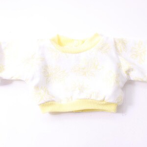 Könnte beinhalten: Ein weißes Sweatshirt mit gelbem Blumenmuster und kurzen Ärmeln. Die Bündchen, der Halsausschnitt und der Saum sind einfarbig gelb. Das Kleidungsstück ist aus einem weichen, leichten Material gefertigt.