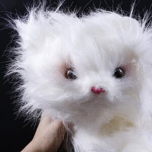 May include: A fluffy white stuffed animal with a pink nose and dark eyes. The toy has long, soft fur and is held against a black background. The toy's fur is slightly disheveled, giving it a playful appearance.