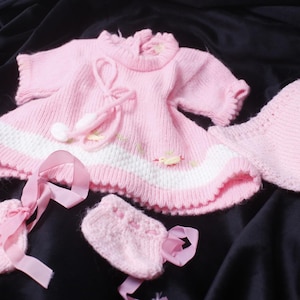 Könnte beinhalten: Ein hellrosa gestricktes Set bestehend aus einem kurzärmeligen Kleid mit einem weißen horizontalen Streifen und kleinen gelben Akzenten, einer passenden Pumphose sowie zwei kleinen gestrickten Schühchen mit rosa Schleifen und Bommeln.
