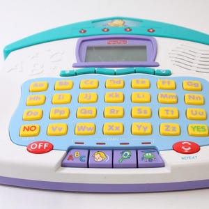 May include: A Fisher-Price ABC learning toy in white, teal, purple, and yellow. The toy features a keyboard with lettered keys, a small screen, and buttons labeled "OFF", "YES", and "REPEAT". The toy is designed for early childhood education.