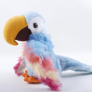 May include: A plush parrot toy featuring a soft light blue body, a large yellow beak, and a dark brown base. The wings display a gradient of pink, yellow, and light blue faux fur. It has white patches around its black eyes and orange feet.