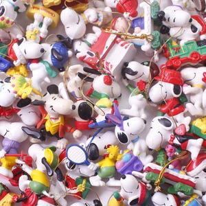 May include: A collection of Snoopy character ornaments in various poses and outfits. The ornaments are white with black accents, and feature Snoopy in different costumes, including a Santa hat, a chef's hat, and a red jacket. Some have gold string for hanging.