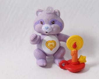 May include: A lavender Care Bear-like figure with purple accents around the eyes and a heart-shaped lightbulb on its belly. It stands next to a red candle holder with a yellow candle and a star.