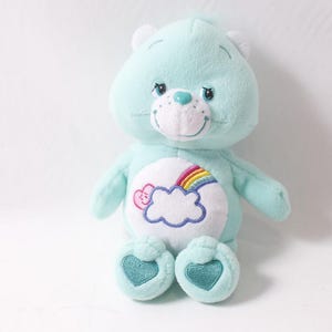 May include: A light blue plush bear with a white belly featuring a rainbow and cloud design. The bear has a smiling face, heart-shaped feet, and white accents on its ears. A classic children's toy.