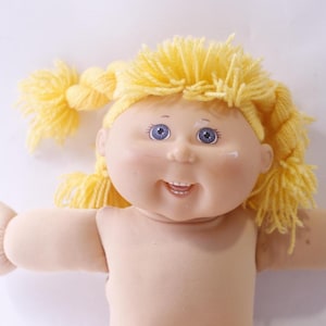 May include: A doll with a soft, light-toned fabric body and head, featuring bright yellow yarn hair styled in two side braids. The doll has blue eyes, a small nose, and a wide, smiling mouth showing two small white teeth on the lower jaw.