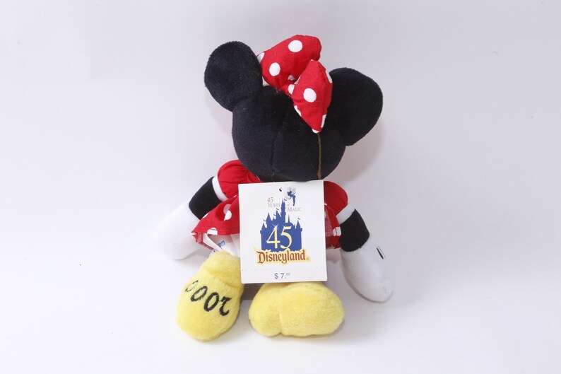 May include: A red and white polka dot plush Minnie Mouse toy wearing a red bow with white polka dots. The toy is holding a tag that says "45 Disneyland $7.99". The toy's feet are yellow and have the number "2000" written on them.