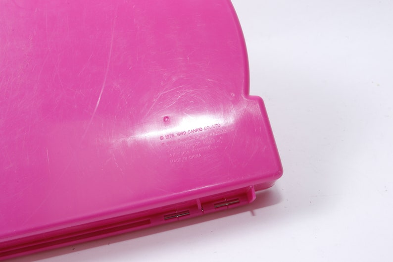 May include: A pink plastic case with the text "&copy; 1976, 1999 SANRIO CO., LTD. ALL RIGHTS RESERVED. MADE IN CHINA" printed on the bottom.