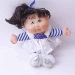 May include: A Cabbage Patch Kid doll with black hair, green eyes, and a white dress with blue and white striped sleeves. The doll is wearing silver shoes and has a white star on its cheek.