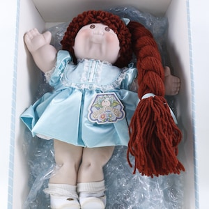 Puede incluir: Una muñeca Cabbage Patch Kids con pelo de lana roja en un vestido de satén azul con ribete de encaje blanco. La muñeca lleva zapatos blancos y está sentada en una caja blanca con forro estampado azul y blanco.