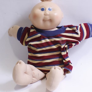 Könnte beinhalten: Eine Cabbage Patch Kid Puppe, die ein gestreiftes Shirt mit roten, gelben und blauen Streifen trägt. Die Puppe hat einen kahlen Kopf mit blauen Augen und einen rosigen Teint.