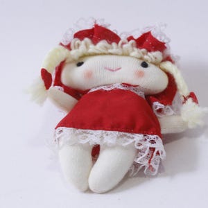 May include: A small, handmade doll with a red dress and white lace trim. The doll has blonde hair and a white face with rosy cheeks.