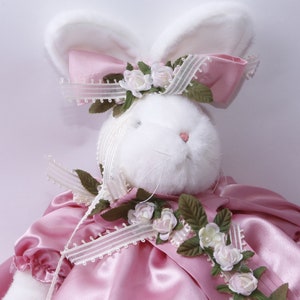 Peut inclure: Un lapin en peluche blanc portant une robe en satin rose avec une bordure en dentelle blanche et un n&oelig;ud rose avec de la dentelle blanche et des accents floraux.