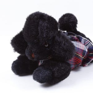 May include: A black plush toy dog with a plaid skirt. The dog has a fluffy, textured appearance and is lying down. The skirt features a red, blue, and white plaid pattern.