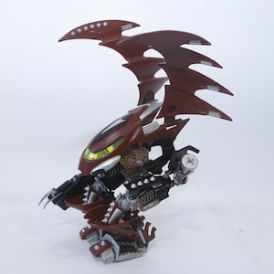Puede incluir: Una figura de acción roja y plateada con alas, que se asemeja a un demonio o una criatura. La figura tiene una cabeza grande con ojos verdes brillantes y un diseño detallado.