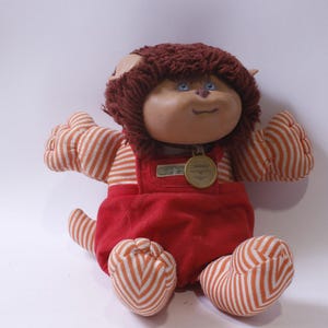 Puede incluir: Un juguete de peluche con pelo marrón, ropa a rayas rojas y blancas y un gorro a rayas rojas y blancas. El juguete tiene una cara redonda con ojos azules y una pequeña nariz redonda. El juguete lleva un mono rojo con una etiqueta dorada que dice "Cabbage Patch Kids".
