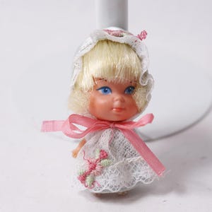 May include: A small doll with blonde hair and blue eyes, wearing a white lace dress with pink and green embroidery. The doll has a pink ribbon tied around its neck and a white bonnet.