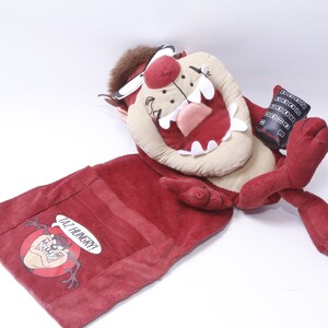 May include: Red plush toy of Taz the Tasmanian Devil from Looney Tunes. The toy is lying down with its mouth open and tongue sticking out. It has a pocket on its back that says "I'm Hungry!"
